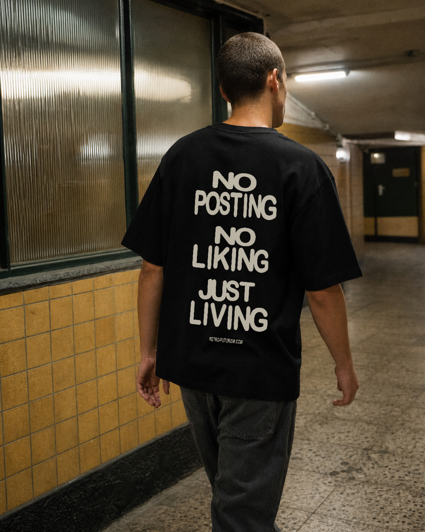 Three-quarter rear view of a model walking through a worn tiled transit concourse in a black Just Living Tee with the bone-colored phrases NO POSTING, NO LIKING, and JUST LIVING arranged vertically across the back.