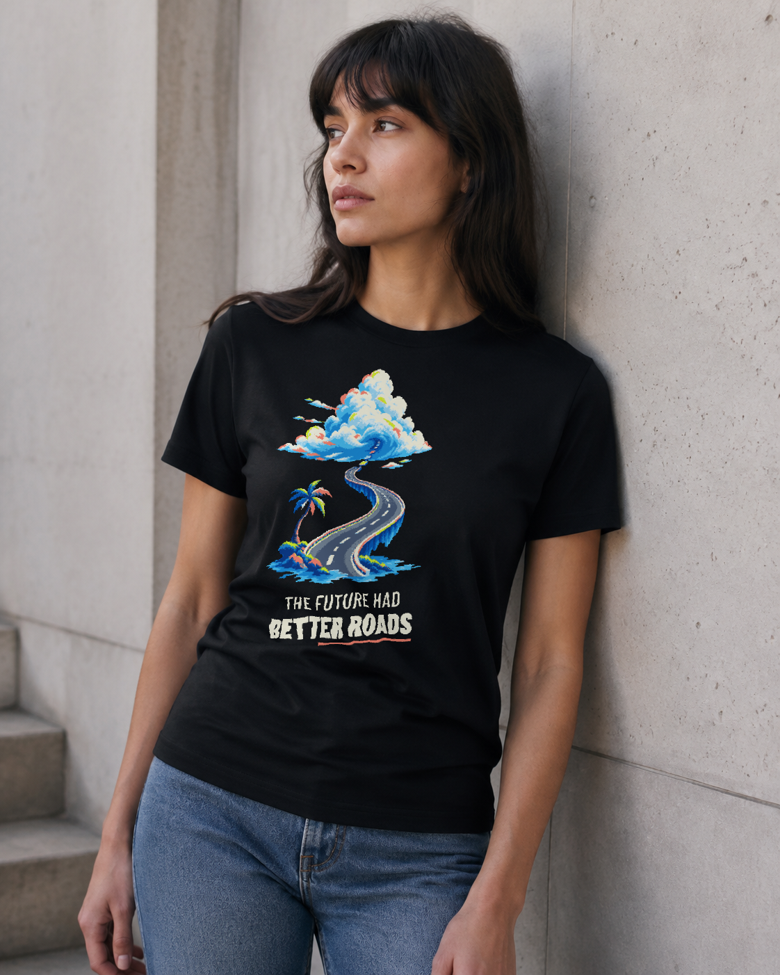 Relaxed leaning view of a model wearing the Black The Future Had Better Roads Tee with the pixel-art coastal-road graphic on the front.