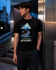 Off-axis standing view of a model wearing the Black The Future Had Better Roads Tee with the pixel-art coastal-road graphic on the front.